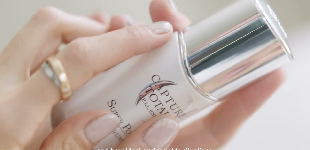 Hands hold a silver serum bottle with a dropper over a light stone surface. Frame from the Dior Capture Totale beauty film.