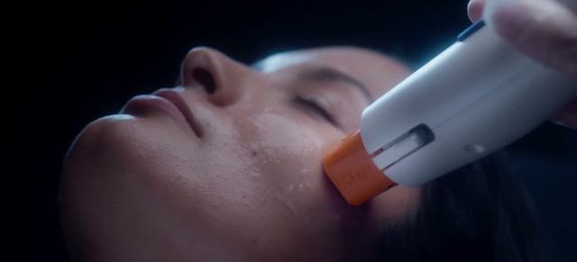 A white handheld device with an orange tip rests on a cheek of a person with eyes closed. Frame from the commercial Saelo, Skin treatments.